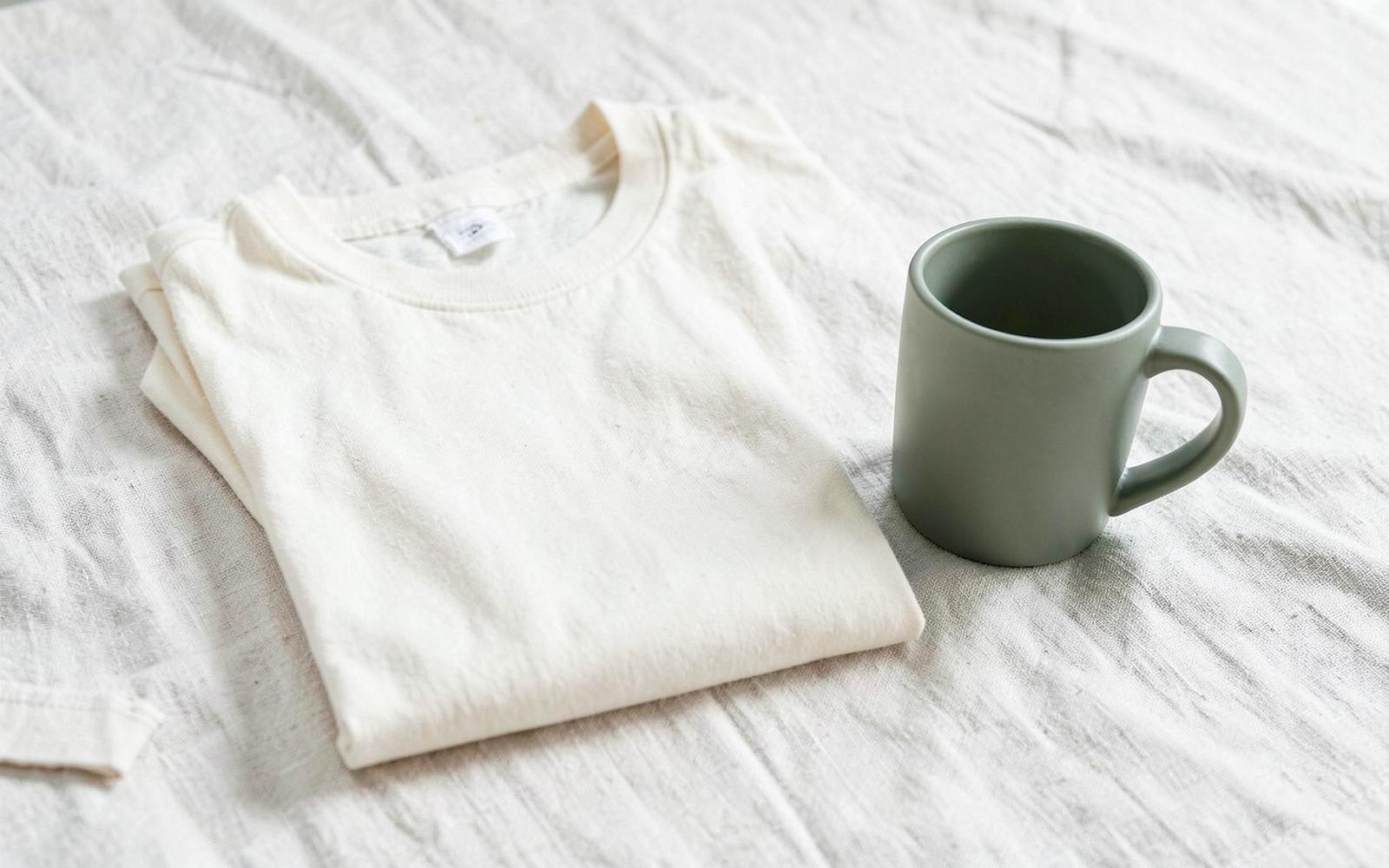 A folded cream t-shirt beside a sage green ceramic mug on linen